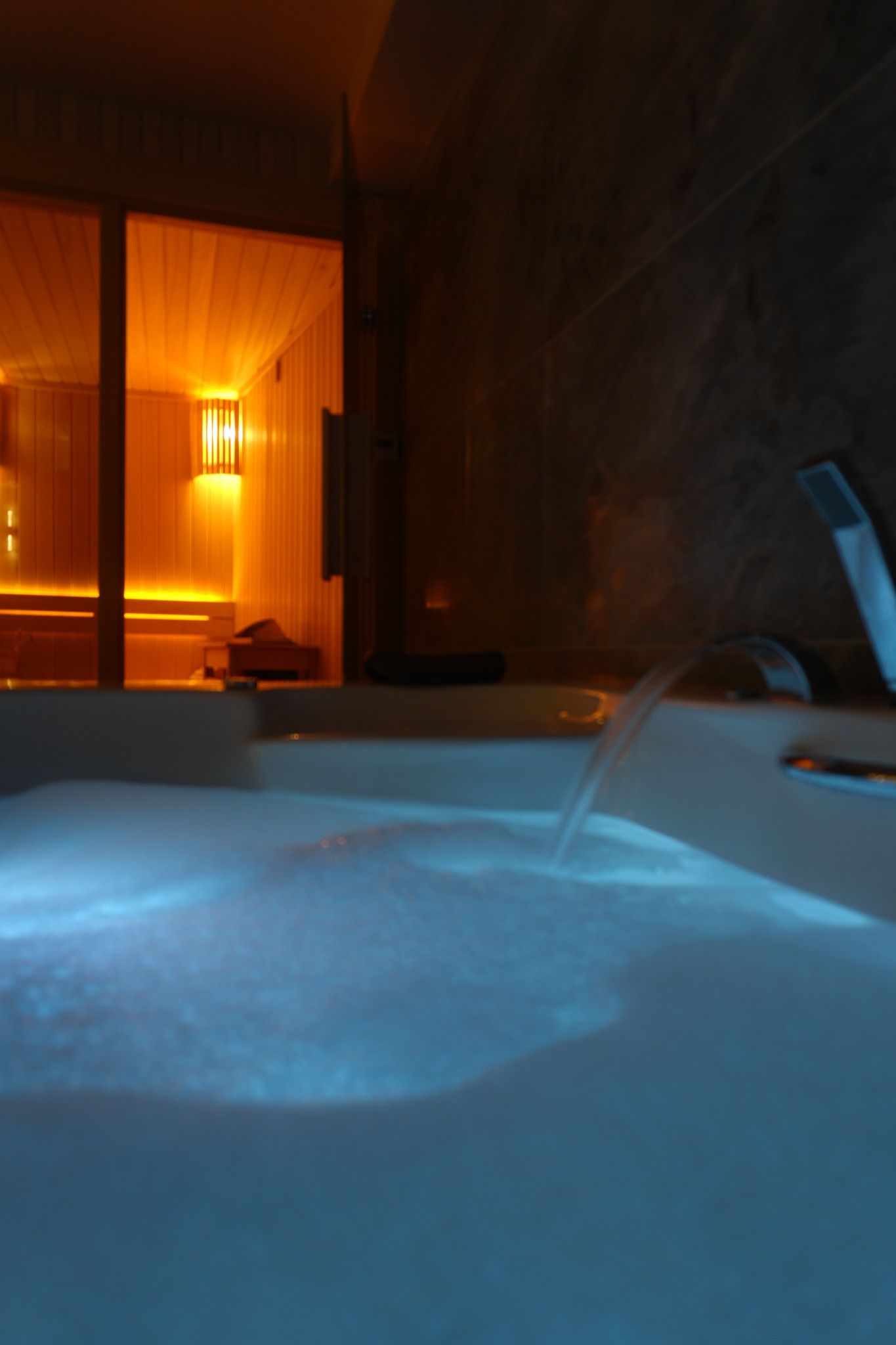 Loş jakuzi alanında, mavi ışıklı suya akan çeşme; arkada sauna ışığı.
